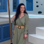 Smiling woman in an olive-green button-down dress with a beige belt, standing by a white staircase and blue windows.